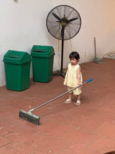 In Cổ Lũng Commune, Vietnam, a young child, appearing to be around 2-3 years old, stands on a reddish-brown tiled floor, holding a floor squeegee with a blue handle. The child is wearing a light yellow ruffled dress and sandals. Behind the child, a large black oscillating fan with a yellow cord is mounted on a stand. To the left of the fan are two large green trash cans with lids. Further in the background, a dustpan and broom are propped against a wall. The overall environment suggests an outdoor or semi-outdoor space with a tiled floor and a simple, functional setting.