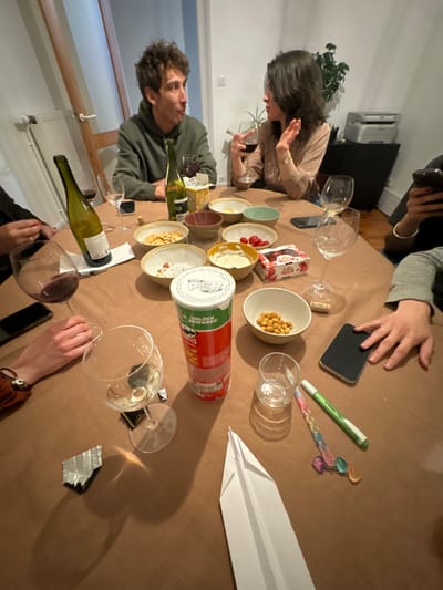 The scene depicts an indoor social gathering around a brown-covered table in Roanne, France. Four individuals are partially visible. A male with curly brown hair, wearing a green hooded sweatshirt, is seated, looking towards a female and appearing to be mid-conversation. The female, with dark, shoulder-length hair, wears a light-colored top, holds a red wine glass, and gestures with her right hand while speaking. Two other individuals' hands and forearms are visible on the left and right sides of the table, each with a smartphone nearby.

The table is laden with multiple items:
*   Several wine glasses, containing red wine, traces of white wine, or empty.
*   Two empty wine bottles, one green and one amber.
*   Numerous small bowls, some containing snack remnants (chips/crackers), one filled with cherry tomatoes, another with a white dip, and one with chickpeas/nuts.
*   A red and white Pringles can (Original flavor).
*   A folded white paper airplane.
*   A crumpled piece of silver foil.
*   A transparent short glass, a wine cork, a green marker pen, and a colorful beaded pen/stylus.
*   A small white snack box with red text (e.g., "AriZona").

The background features a light-colored wall, a white-framed window with a wooden object leaning against the wall, and a white radiator. Further back, a desk or shelf holds a printer/scanner and a potted plant. The lighting suggests an indoor evening setting.