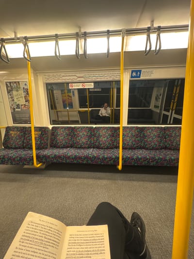 An interior view of a Perth public transport train carriage, likely a Transperth train, shows the reflection of the surrounding environment in the windows. The seats are upholstered in a dark fabric with a repeating pattern of red, green, and blue motifs. Yellow poles, commonly found in public transport for passengers to hold onto, are visible. In the foreground, an open book rests on the lap of a person wearing black pants and a black sneaker. The book's pages contain text, with some passages highlighted. Through the windows, the reflection shows other passengers, one individual in particular is seated and looking at their phone. Another section of the window displays signage indicating "NO SMOKING," "NO VAPING," and "NO DRINKING." A sign on the seats reads "PRIORITY SEAT." The overall environment suggests a typical commute on public transportation.
