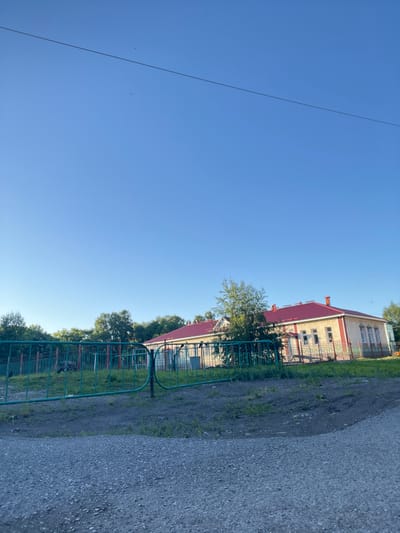 This image depicts a residential building with a red roof, possibly a school or administrative building, in Kiselyovsk, Russia. The building is light-colored with red trim and has multiple windows. It is partially obscured by a green fence. To the left of the building, there is a gravel pathway and some green vegetation. The sky is a clear, bright blue, suggesting a sunny day with good weather. The sun appears to be high in the sky, indicating it is likely midday. There is a thin black wire stretching across the sky. No people are visible in the image. The overall impression is one of an ordinary day in a semi-urban or rural setting.