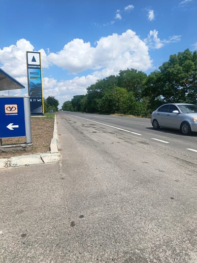 The image depicts a roadside scene in the city of Koshary, Ukraine, on a clear day with scattered clouds. A paved road runs through the frame, with a silver sedan driving on the right lane. To the left of the road, a blue sign with a white arrow and the "UKRNAFTA" logo indicates a direction. A tall, yellow sign also displays the "UKRNAFTA" logo, along with information about "ZAPRAVLYAYU" (refueling), "ENERGY," and a "WC" symbol. Trees line the right side of the road, forming a dense backdrop. The foreground shows a concrete curb and a dirt patch with sparse vegetation. The scene is typical of a rural or suburban area with a gas station.