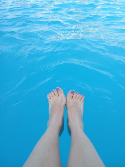 The image shows a person's legs and feet submerged in bright blue water. The water has a rippled texture, suggesting movement or light play on its surface. The feet are positioned in the lower center of the frame, with the toes pointing upwards. The toenails are painted a vibrant blue color. The skin on the legs and feet appears fair and has a slightly textured appearance, with some subtle markings. The water level comes up to about the mid-calf area. The scene is dominated by the expansive blue of the water, creating a sense of depth and possibly serenity. The image is shot from a top-down perspective, looking directly at the legs and feet. There is no visible text in the image. The overall impression is one of relaxation and enjoyment of the water.