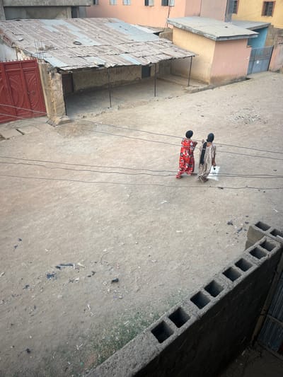 An aerial view shows two individuals walking on a dirt road. The person closer to the camera wears a vibrant red patterned dress, while the other dons a light-colored outfit. Power lines traverse above them. To the left, buildings with metal roofs and red doors stand tall. A concrete structure with square openings lines the upper right corner, casting shadows. The overall scene depicts a simple street view in what appears to be a developing urban area.
