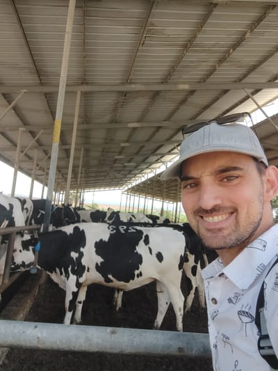 A man, wearing a light blue baseball cap and a white shirt with line drawings of various objects, is taking a selfie in a barn. He is smiling broadly at the camera. Behind him, several black and white cows are visible in stalls. The barn has a corrugated metal roof supported by metal beams. The ground appears to be dirt or mud. The image is taken from a slightly low angle. The location is identified as a farm in the Yoav Regional Council, Israel.