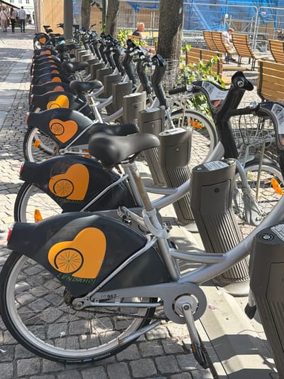 The image shows a row of parked bicycles for rent in Lund, Sweden. The bicycles are silver with black fenders featuring an orange heart logo and the text "LUNDAHOJ". The bikes are arranged in a line on a cobblestone surface. In the background, there are wooden benches and a blue construction barrier. Several people are visible, including a man with blond hair sitting on a bench and other individuals walking in the distance. The scene appears to be a sunny day in a public outdoor area.