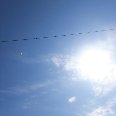 The scene, located in Gimpo-si, South Korea, captures an overhead view of a bright blue sky. A brilliant sun occupies the upper right quadrant, emitting significant glare and a broad white lens flare. Scattered, thin white clouds are present, primarily in the lower right and mid-left areas. A single dark, segmented wire spans horizontally across the mid-section of the sky. A distinct small, circular, yellowish-orange lens artifact is visible in the mid-left portion of the frame. No individuals, ground structures, or specific activities are depicted.