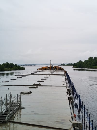 The image shows a cargo ship on the Svir River in Vytegra, Russia. The ship is loaded with yellow cargo containers and is being towed by a tugboat. The sky is overcast and the water is calm. The image is taken from the deck of the cargo ship, looking towards the front of the ship. There are no people visible in the image.