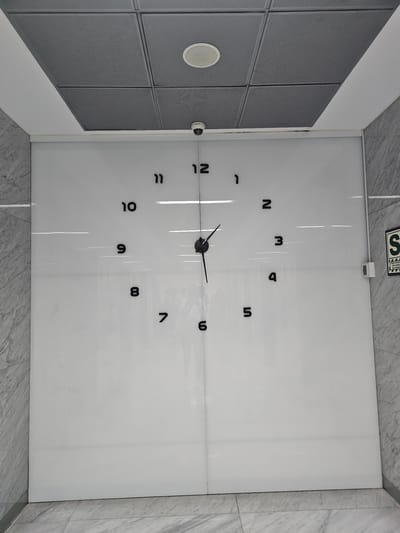 The image shows a large clock mounted on a white wall, likely in an indoor location within the city of La Molina, Peru. The clock face is minimalist with black numerals displayed against the white background. The hands indicate a time around 11:23. The walls adjacent to the clock wall have a gray, patterned surface. Part of a ceiling with a circular light fixture is visible, indicating an interior space. A small sign is visible in the upper left corner with the letter "S" and some illegible text, which is a seismic safety symbol.