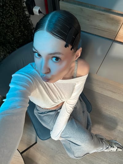 A young person is taking a selfie in what appears to be a home setting in Zakharovo, Russia. The person is sitting on a chair, with their legs extended and wearing grey sweatpants and a white long-sleeved shirt. Their hair is neatly slicked back with several clips in place, suggesting they might be in the process of styling it. They are also wearing athletic shoes. The background shows a wooden-paneled wall and a grey floor. The lighting in the image is cool-toned, possibly from artificial sources, casting a blueish hue on the subject and their surroundings. The person's expression is somewhat pensive as they look directly at the camera. No specific time of day or weather conditions can be determined from the image. There is no visible text in the media file.