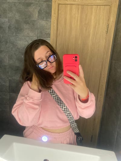 A young woman with dark brown hair and dark-rimmed glasses is taking a mirror selfie. She is wearing a pink cropped sweatshirt and matching pink sweatpants. A black crossbody bag with a geometric pattern is slung over her shoulder. She is holding a red smartphone in her right hand. The background shows a tiled wall and a wooden door. The scene appears to be a bathroom, possibly in a private residence. There is no discernible information within the image that places the location in Chaikovsky, Russia.