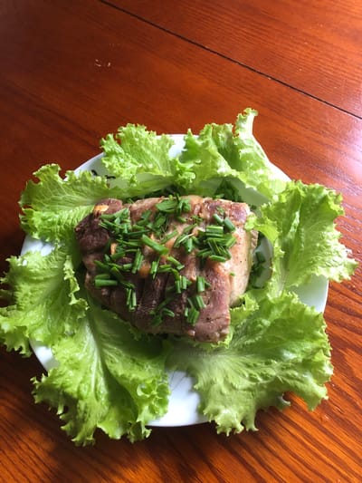 A close-up, overhead shot shows a piece of cooked meat, garnished with chopped green onions and possibly garlic slivers, resting on a bed of vibrant green lettuce leaves. The plate is white with a subtle pattern around the rim and sits on a polished wooden table. The lighting suggests an indoor setting, likely a home dining area. There are no people visible in the image, and the focus is solely on the food presentation. The location is not specified within the image itself, and therefore, it cannot be stated to be in Barrie, Canada.