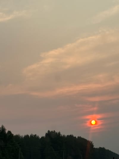 This image captures a sunset over Staryi Poros, Russia. The sun, a bright orange orb, is low in the sky, partially obscured by wispy clouds that are tinged with pink and yellow. A dark silhouette of trees forms the horizon line below, with power lines strung across the foreground. The overall atmosphere is serene and dusky, typical of a sunset.