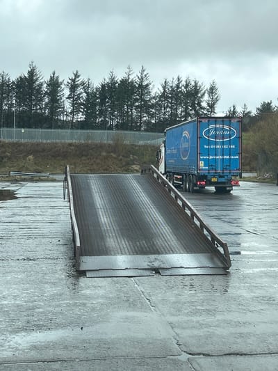 The image, likely taken in Shotts, United Kingdom, shows a lorry with "Jordan" branding backed up to a loading dock or platform. The lorry is blue and has the phone number 01505 152166 and website www.jordanltd.co.uk displayed on its rear doors. The loading platform is made of metal grating with a metal railing. The ground is wet and appears to be concrete. A line of trees and a fence are in the background. The sky is overcast.