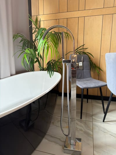 A freestanding bathtub faucet with a handheld shower attachment is positioned in the foreground of a bathroom. The faucet is made of chrome and features a curved spout. Behind the faucet, a lush green potted plant is visible. To the right, a grey upholstered chair with black legs is present, with another chair partially visible to its right. The background consists of a wall with vertical wooden panels and black trim. The floor is tiled with a marble-like pattern. The scene appears to be indoors, in a modern bathroom setting. There is no indication of the specific location within the city of Семилуки, Russia.
