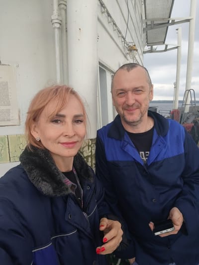 Two people, a woman and a man, are posing for a selfie on what appears to be the deck of a ship. The woman is in the foreground on the left, with short, light-colored hair and a warm smile. She is wearing a dark blue winter coat with a gray faux fur collar. Her left hand is resting on her coat, while her right hand is reaching out, holding a cell phone. The man is standing to her right, also smiling. He has short graying hair and a beard. He is wearing a dark blue and royal blue work jacket over a black t-shirt.

The background shows the exterior of a boat or ship, with white walls and metal piping. The sky is overcast, suggesting a cloudy day. In the distance, a body of water and what looks like the shore can be seen, along with some industrial elements and boat rigging. The scene seems to be taking place in a working environment, possibly a port or waterway, given the context of Novaya Svobodka, Russia. The overall mood is casual and friendly, indicated by the smiles of the subjects. There is some faded text on a white panel behind the woman, but it is largely illegible.