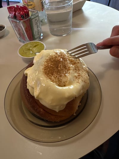 A cinnamon roll covered in thick, white icing and sprinkled with cinnamon sits on a brown glass plate. A silver fork is being used to cut into the roll. To the left of the plate is a glass of water and a container holding packets of Splenda. A small cup containing a green sauce is also visible. The scene is set on a light-colored table.