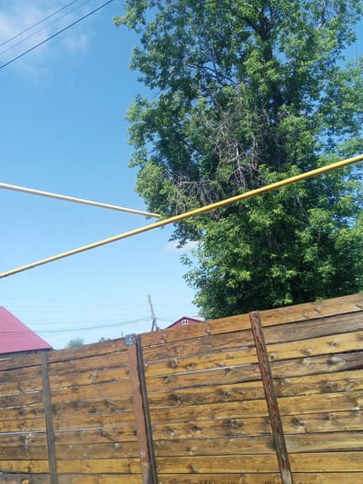 This image depicts a rustic outdoor scene, likely in Maslyanino, Russia, characterized by a wooden fence in the foreground and a large, leafy tree against a bright blue sky in the background. The fence, constructed of horizontal wooden planks, appears weathered and has a reddish-brown hue. Two vertical wooden posts support the fence, and one of these posts is partially covered by a reddish, possibly metal, pipe.
In the upper portion of the image, two parallel yellow pipes are visible, running horizontally across the frame. Beyond the fence and the tree, a portion of a red-roofed building is discernible on the left, with a smaller building featuring white window frames visible on the right, further in the distance. Electrical wires are also present, strung across the sky. The overall impression is of a somewhat rural or suburban setting, possibly a backyard or a public space with utility infrastructure.