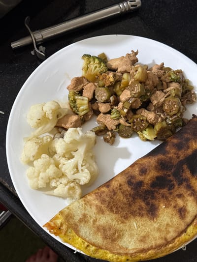 A top-down shot depicts a white plate containing what appears to be a meal. The dish comprises three distinct sections: one with sauteed chicken, broccoli, and okra; another with cooked cauliflower florets; and the third with a partially burnt, folded flatbread, potentially a dosa. Adjacent to the plate is a long, silver kitchen lighter. No people are directly visible in the frame, but the edge of a foot is visible to the right. The setting appears to be indoors, likely a domestic kitchen or dining area, based on the proximity to a food preparation surface. There is no evidence to directly confirm the location as Marra, India.