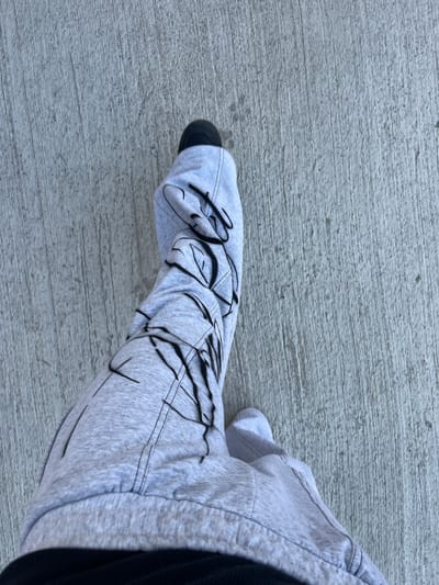 The image shows a person's foot and lower leg clad in light grey sweatpants with black graffiti-style designs. The pants are cuffed at the ankle, revealing a black sock and the toe of a shoe. The person is standing on a concrete surface, possibly a sidewalk. Due to the lack of contextual information and visual landmarks, it is impossible to confirm the location as Bahía Blanca, Argentina.