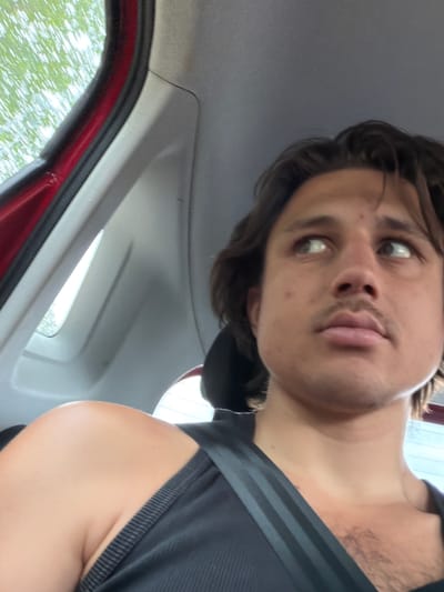 This is a first-person, low-angle selfie taken from inside a car. The subject is a young man with brown hair and light eyes, looking directly at the camera with a slightly surprised or curious expression. He is wearing a dark tank top with thin grey stripes on the shoulder strap. The background shows the interior of the car, including the headliner and a portion of the red door frame, with a blurred view of green foliage through the window, suggesting it's daytime and likely spring or summer. The image appears to be taken in Stammham, Germany. There is no visible text in the image.