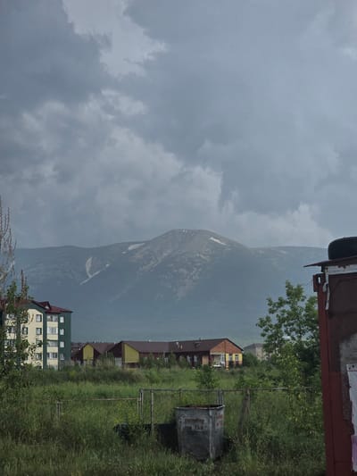 The image shows a cloudy sky over a mountainous landscape. In the foreground, there are several buildings, including a tall, light-colored apartment building with green accents, and several smaller, lower buildings with yellow and red facades. There is a lot of green grass and vegetation in front of the buildings, with a metal fence and a trash bin visible. To the far right, a portion of a dark-colored structure with a tire on top is visible. The scene appears to be a residential area near a mountain range. The location is identified as the city of Ridder, Kazakhstan.