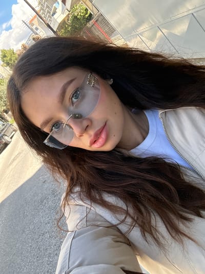 A young woman is photographed outdoors. She has long, brown hair, fair skin, and is wearing sunglasses with a logo visible on one lens. Her attire includes a white t-shirt and a light-colored jacket. The background shows an urban environment, including a street and buildings. The location is within the Municipal Unit of Elliniko, Greece.