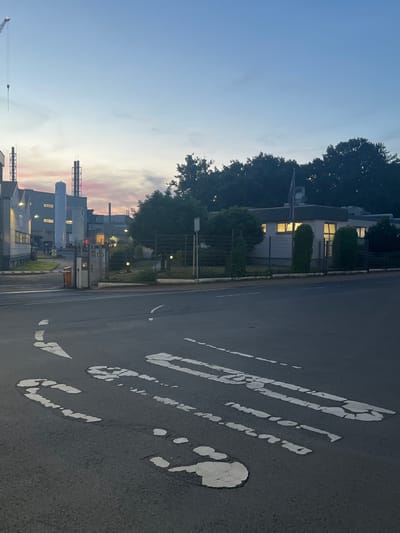 The image depicts the entrance to an industrial complex in Stadtallendorf, Germany, during dusk. In the foreground, a paved road shows faded white lane markings, including what appears to be a pedestrian crossing. To the left of the road, a metal barrier gate is partially open, with a small attendant booth beside it. The industrial complex itself features several large, white cylindrical tanks and other industrial buildings with illuminated windows. In the background, silhouetted trees form a dark line against the twilight sky, which is painted with soft hues of pink and blue from the setting sun. A tall construction crane is visible in the far left of the image. The scene suggests the end of a workday, with the facility still partially lit and active.