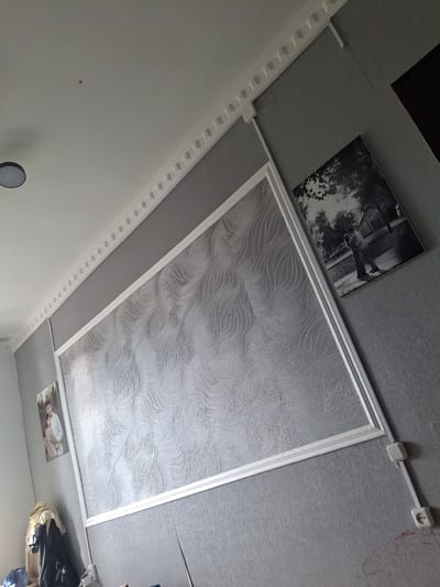 The image depicts an interior view of a room with grey textured walls and a white ceiling. A white decorative molding runs along the ceiling-wall junction. A large framed panel with a swirling silver pattern is mounted on the wall. To the right of this panel is a framed black and white photograph of a couple embracing outdoors. On the left side of the large panel, a smaller framed color photograph shows a couple standing close. The room features electrical outlets and wiring visible on the wall, some contained within white conduits. A ceiling light fixture is partially visible. A light fixture is seen on the ceiling, and a small dark spot is also present on the ceiling. A dark object, possibly a piece of furniture or an appliance, is visible in the upper right corner. Clothing items are draped on a coat rack in the lower left. The overall impression is of a lived-in domestic space. The location is stated as Мичурин ауылдық округі, Kazakhstan.