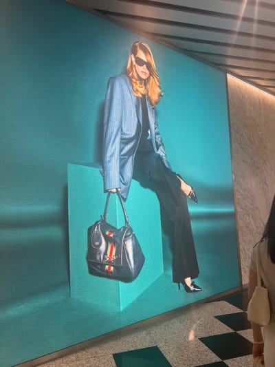 The image displays a large advertisement on a wall, featuring a model in a pinstripe blazer, black pants, sunglasses, and black heels. She is seated on a teal-colored cube and holding a dark blue handbag with red and green stripes. To the right of the advertisement, a woman with long dark hair, wearing a light-colored t-shirt and carrying a cream-colored bag, is walking past. The advertisement is illuminated by ceiling lights, casting shadows on the tiled floor, which has a checkerboard pattern of dark and light tiles. The environment suggests an indoor public space, likely a shopping mall or a hotel lobby, possibly in Guangzhou City, China, given the presence of the Gucci advertisement and the architectural style often seen in modern commercial areas of major Chinese cities.