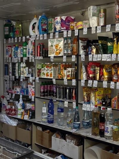 The image depicts a well-stocked retail shelf, seemingly inside a store in Слобода, Russia, based on the Russian labeling on many of the products. The shelves contain a variety of products, including bottled water, juice, coffee, cleaning supplies, snacks, and paper products. Price tags are visible next to most items, indicating retail sale. The shelves are metal and appear to be arranged vertically. The image is taken from an angle, with the shelves slightly tilted. There are cardboard boxes stacked near the shelving unit.