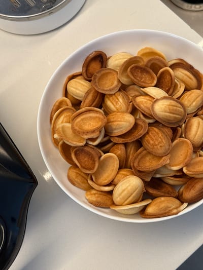 A top-down forensic view captures a white, oval-shaped bowl overflowing with numerous baked pastry shells, commonly known as "Oreshki" or "nut cookies," situated on a light-toned, possibly off-white, kitchen countertop in Башкултаево, Russia. The cookies exhibit varying shades of golden-brown to deep amber, indicating differential baking, with some displaying distinct ridged patterns consistent with a molded preparation. The majority are hollow shells, while some appear to have two halves joined, creating a complete "nut" shape. To the upper left, a portion of a silver-toned kitchen appliance with a slotted top and a white base is partially visible. In the lower left quadrant, a segment of a dark, possibly black, object with a smooth, reflective, curved surface is present. The countertop exhibits subtle reflections from ambient overhead lighting. No individuals are visible.
