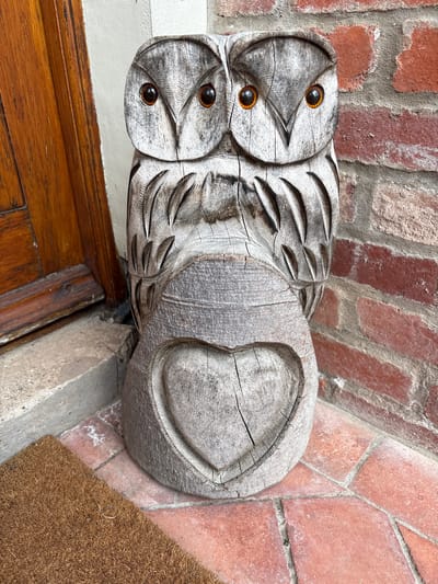 A wooden sculpture of two owls is placed near a doorway. The sculpture has detailed carvings of feathers and distinct, rounded eyes on each owl. The wood appears weathered, with visible cracks. The sculpture rests on a concrete step next to a wooden doorframe and a doormat. A brick wall is visible in the background. The location is identified as Minsterley, United Kingdom.