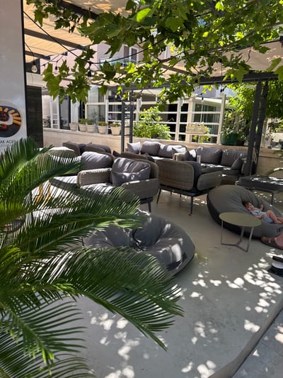 The image depicts an outdoor seating area, likely a cafe or restaurant patio, in Makarska, Croatia. The area is furnished with a variety of comfortable seating options, including plush grey beanbag chairs and wicker-framed sofas with grey cushions. A small child is visible lounging on one of the beanbag chairs, looking towards the camera. Lush green foliage from overhead trees casts dappled shadows on the concrete floor, suggesting a sunny day. In the background, a building with large windows and potted plants indicates an urban setting. The overall impression is one of a relaxed and inviting outdoor space.