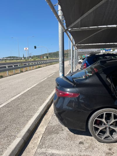 The image shows a black car parked under a covered parking area with a mesh shade. The car is facing towards the right of the frame, with its rear visible. To the left of the parking area, there is a road with a guardrail and more parking spaces visible in the distance. In the background, there are hills with vegetation and a clear blue sky. A sign for "AVIN" gasoline is visible in the distance. The scene appears to be a parking lot near a highway or major road, possibly at a service station or rest stop, in the Municipal Unit of Iera Poli Messologhiou, Greece.