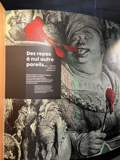 This image displays a large, brightly lit exhibition panel located within the Cité internationale de la gastronomie et du vin in Dijon, France. The panel features an intricate, black-and-white illustration in an engraving style, with specific elements highlighted in red.
The central subject on the right is an extremely corpulent man depicted in profile, facing left. He wears a large, elaborate turban and an ornate, heavy-linked necklace or collar. His eyes are rolled back, and his tongue extends from his mouth, suggesting intense sensory pleasure or gluttony. He holds a thin, long-handled utensil, possibly a spoon or small fork, with a vibrant red, dripping substance at its tip, positioned near his mouth. His left hand, largely obscured, appears to hold a bowl or container from which the substance was taken.
In the upper left quadrant, a group of smaller, less defined figures observe the scene. One figure, centrally positioned within this group, is entirely highlighted in red and appears to be offering or presenting two red spoons.
The lower left section of the panel shows two additional figures, also in the black-and-white engraving style. One stands upright, looking right, while the other, with its back partially to the viewer, is a grotesque or heavily cloaked figure holding a large, rounded object resembling a pot or barrel.
Overlaid on the illustration, primarily in the left and center, is white text. The most prominent title reads "Des repas à nul autre pareils..." followed by "Meals beyond compare" in English. Below this, a French paragraph describes "Haughty guardians of changing traditions, connoisseurs who like to stuff themselves... the French have the best cuisine in the world and always a good reason to bend an elbow!" with an accompanying English translation. The overall theme conveyed is a celebration of French gastronomy, abundance, and culinary pride.