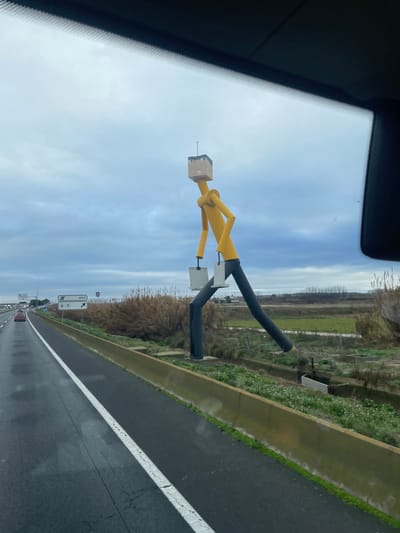 The image, captured from the interior of a vehicle, displays a large, stylized anthropomorphic sculpture situated roadside along the A-2 highway near Bell-lloc d'Urgell, Spain. The sculpture, approximately three stories tall, features a beige, rectangular head with minimal black facial features and a thin vertical antenna. Its body consists of a yellow, cylindrical torso and arms, with two rectangular, light grey objects (resembling bags or boxes) held in each hand. The figure's long, slender legs are dark grey/black, posed in a dynamic walking stride, with the left leg extended forward and the right leg bent, partially obscured.

The foreground depicts a damp, asphalt highway with a distinct white solid line marking the shoulder and a dashed white line for the traffic lane. A low, light-colored concrete barrier separates the highway from the narrow grassy strip where the sculpture stands on a small concrete base. Beyond this, a shallow ditch or canal with visible water runs parallel to the road. The background is dominated by a field of tall, dry, straw-colored reeds and grasses, with a line of dark trees in the far distance. Overcast, light grey clouds fill the sky. In the far background, a red passenger vehicle is visible on the highway, and white directional road signs displaying symbols and numbers like "A-2" and "480" are present. No human individuals are visible in the scene.
