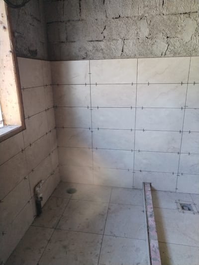 The image depicts an interior view of a room undergoing construction or renovation, specifically a tiled corner. The walls are partially tiled with light-colored square tiles, marked with horizontal and vertical black lines and small cross-shaped tile spacers. The top portion of the walls appears to be rough concrete block. A wooden window frame is visible on the left side. In the corner, a white pipe extends downwards, and the floor is also tiled with similar square tiles. A dark circular drain is visible on the floor, and further to the right, a square drain fixture. A pink and grey metal bar lies diagonally across the tiled floor. The overall environment suggests a bathroom or shower area in a state of unfinished construction. There are no people visible in the image.