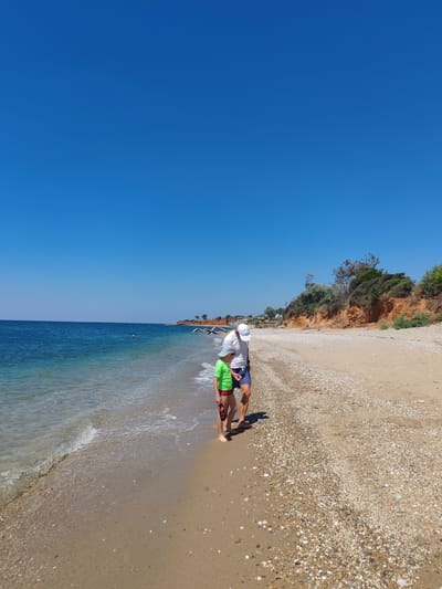 A father and son stand on a pebble and sand beach in Alexandroupolis, Greece, near the Aegean Sea. The father, wearing a white t-shirt, white cap, and blue swim shorts, looks down at his son. The son, dressed in a green t-shirt and red swim shorts, holds a small object in his hand. Gentle waves lap the shore under a clear blue sky. In the background, a rugged, vegetated cliff face borders the sea, with scattered driftwood on the beach.