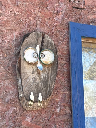 A photograph shows an art installation on a wall in Маза, Russia. A blue window frame is fixed to the top of a wall constructed from composite wood, possibly OSB or chipboard. Below the window frame, a section of a tree trunk or a large branch acts as a display surface. The items displayed on the wood consist of two bottle caps positioned to resemble eyes, a spoon bridging the gap between them, and two hair clips placed on the opposite side of the wood piece, mimicking a mustache.