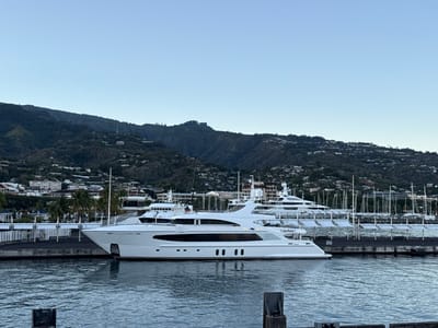 The image displays a harbor scene in Papeete, French Polynesia, likely observed during twilight hours. In the mid-ground, a prominent, large, white luxury yacht named "BIG SKY" is moored alongside a dark-colored dock. The yacht features multiple decks, dark tinted windows, and a sleek hull. Behind and to the right of "BIG SKY," several other white luxury yachts and numerous sailing vessels, identifiable by their visible masts, are docked within the marina. The foreground consists of calm, dark blue-grey water with minimal ripples; a dark, rectangular mooring structure is partially visible in the lower right. Beyond the marina, urban structures and scattered palm trees line the waterfront. The background is dominated by steep, densely vegetated mountains, with numerous residential buildings distributed across their lower slopes. The sky is clear and a pale blue. No individuals, specific actions, or interactions are discernible in the scene.