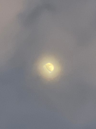 A highly luminous, crescent-shaped object, possibly a celestial body, emits a concentrated yellowish-white light from its core, surrounded by a distinct circular halo of diffused light. This light source is centrally positioned against an entirely uniform light grey sky, suggesting extensive cloud cover or atmospheric haze. No terrestrial features, human subjects, or other discernible objects are present within the frame. The observation occurred in Hale Bandi Harlapura, India.