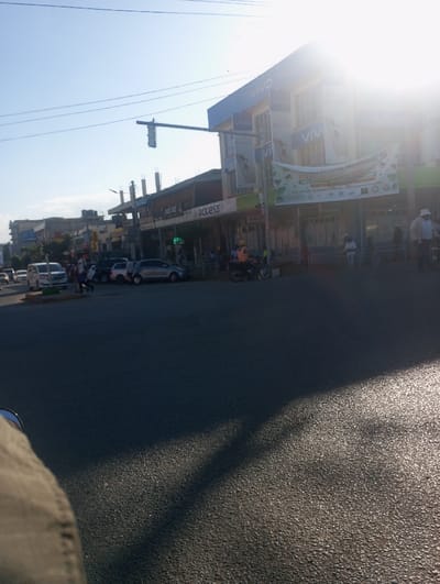 This image depicts a sunlit street scene in Kimumu ward, Kenya, likely during the early morning or late afternoon, given the bright sun causing a significant lens flare on the right side and casting long shadows across the asphalt road.
The scene is bustling with everyday activity. Multi-story buildings line the street, with a prominent blue building on the right heavily branded with "vivo" logos, suggesting a mobile phone store. Below it, another storefront displays an "Access" sign. A large, partly sun-obscured banner hangs from the "vivo" building, appearing to announce a "KIMUMU CULTURAL FESTIVAL."
A traffic light is suspended over an intersection. The street is active with various vehicles, including a white van, several cars, and a motorcycle. Pedestrians are visible walking on sidewalks or standing near storefronts, including a person in an orange vest near the motorcycle and another in a white hat further down the street. Overhead utility lines crisscross the clear blue sky. A partial view of a person's leg, clad in khaki pants, is visible in the bottom-left corner, indicating a low-angle shot, possibly from a vehicle. The image portrays a typical, vibrant commercial street environment.