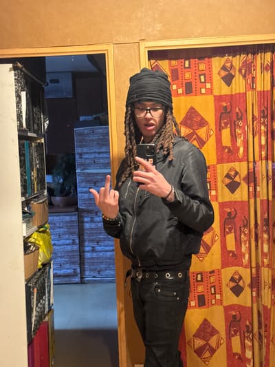 A person wearing glasses, a black head covering, and a dark jacket stands against a backdrop of patterned curtains. They are holding a smartphone and extending their middle finger. The background includes a shelf with various items and a view into a darker room with blue walls. The location is indicated as Orsay, France, though this is not visually verifiable in the image.