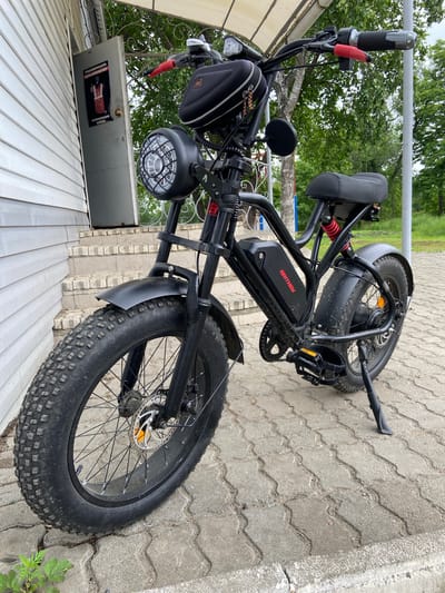 An outdoor, eye-level, full-shot photograph captures a black fat-tire electric bicycle parked on a paved walkway in front of a building with white siding. The bicycle is positioned slightly to the right of the frame. Its prominent features include a large, textured front tire and a matching rear tire, a black seat with red shock absorbers visible beneath it, a black frame, and a black battery pack on the downtube with the red "BRITNEY" logo. The front of the bicycle has a circular headlight with a wire grille. Red grips are on the handlebars, along with a black bag attached. To the left of the bicycle, an open grey door reveals a poster with text and an image of a person in an orange vest. The background consists of trees and a light-colored pole. The paving stones are irregular in shape and color. The scene appears to be a quiet, possibly residential, area. The time of day seems to be daytime, indicated by the natural lighting.