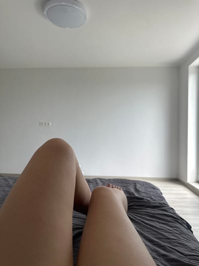 The image depicts a bedroom scene. In the foreground, two legs are visible, lying on a dark gray bed with rumpled sheets. The legs are bare and appear to be resting, with one knee slightly bent and the other leg extended. The skin on the legs shows a subtle texture, possibly goosebumps or minor imperfections.

The background consists of a plain, light gray wall. A ceiling light fixture is visible at the top of the image. On the wall, two white electrical outlets are mounted horizontally. To the right, a large window is present, allowing natural light to enter the room, illuminating the wall and floor. The floor is a light-colored laminate or wood. The overall impression is of a simple, uncluttered domestic space. The location of Velyka Cherniavka, Ukraine, is noted as the setting for this scene.