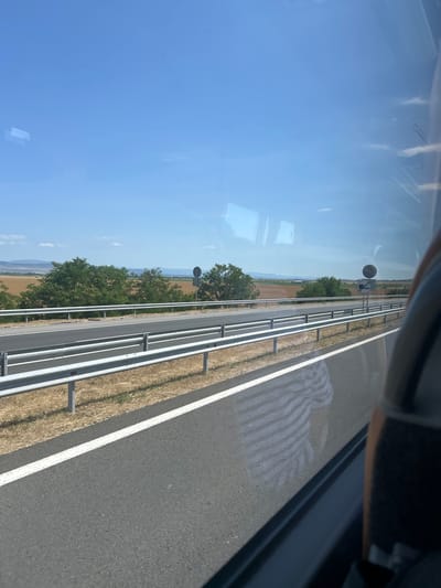 The image shows a view from a vehicle, likely a bus, traveling on a highway. The highway has multiple lanes and is bordered by metal guardrails and dry grass. Beyond the highway, there are fields, trees, and distant hills under a clear blue sky with a few wispy clouds. Two road signs are visible on the right side of the highway. The reflection of a person wearing a striped shirt is visible on the highway surface. The location is identified as Diadovo, Bulgaria.