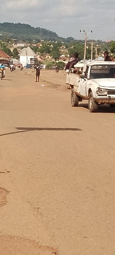 In Gboko, Nigeria, a white pickup truck drives down a dusty, paved road. Several young men are riding in the bed of the truck, some leaning over the sides. One person stands in the middle of the road with their arms raised, looking towards the truck. In the background, buildings and trees are visible, with a large, green hill in the distance. The sky is overcast with clouds. The scene appears to be during the daytime.