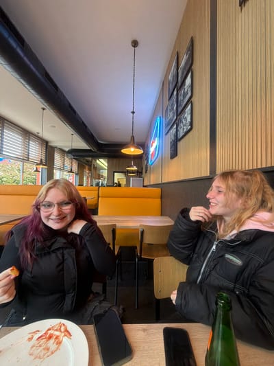 Two young women are seated at a table inside a restaurant. One has blonde hair and is wearing a pink hoodie under a black jacket; she is smiling and looking downward. The other has blonde and purple-tinted hair, wearing glasses and a black jacket, and is holding a food item. A green bottle is visible on the table near the blonde-haired woman. The table also contains a smartphone and a plate with remnants of ketchup. The restaurant has yellow booth seating, wood paneling on the wall, black-and-white framed pictures, and a blue neon sign. The location is presumed to be in Grevenmacher, Luxembourg.