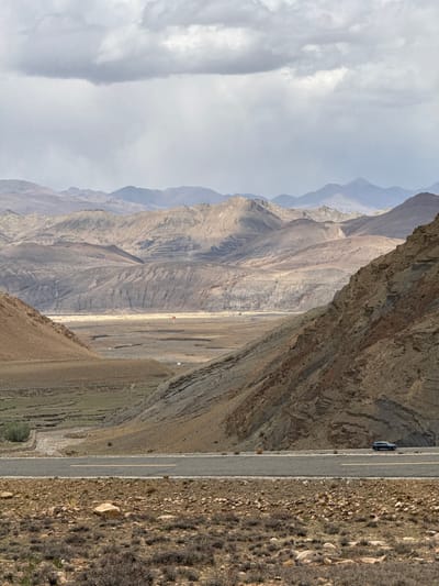 The image depicts a mountainous landscape in Zagor town, China. A paved road runs alongside a steep, rocky cliff face on one side and a dry, barren hillside on the other. A dark-colored vehicle is visible on the road. The background features layers of hazy mountain ranges under a cloudy sky. The vegetation appears sparse and dry.