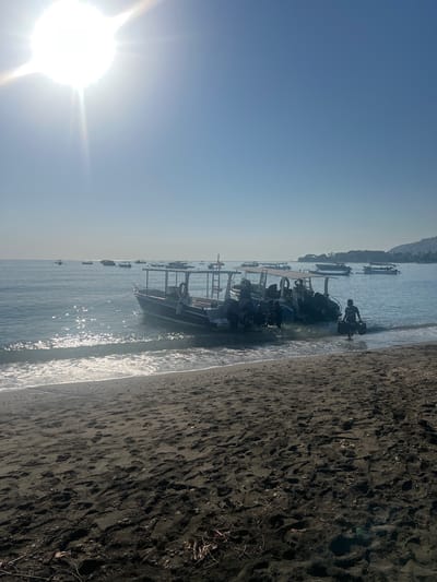 A bright sun shines down on a calm, blue ocean with a clear sky. Several boats are docked near a dark sandy beach, where a person is walking out of the water with two bags. The shore is lined with footprints in the sand. The scene appears to be in the city of Gerokgak, Indonesia.